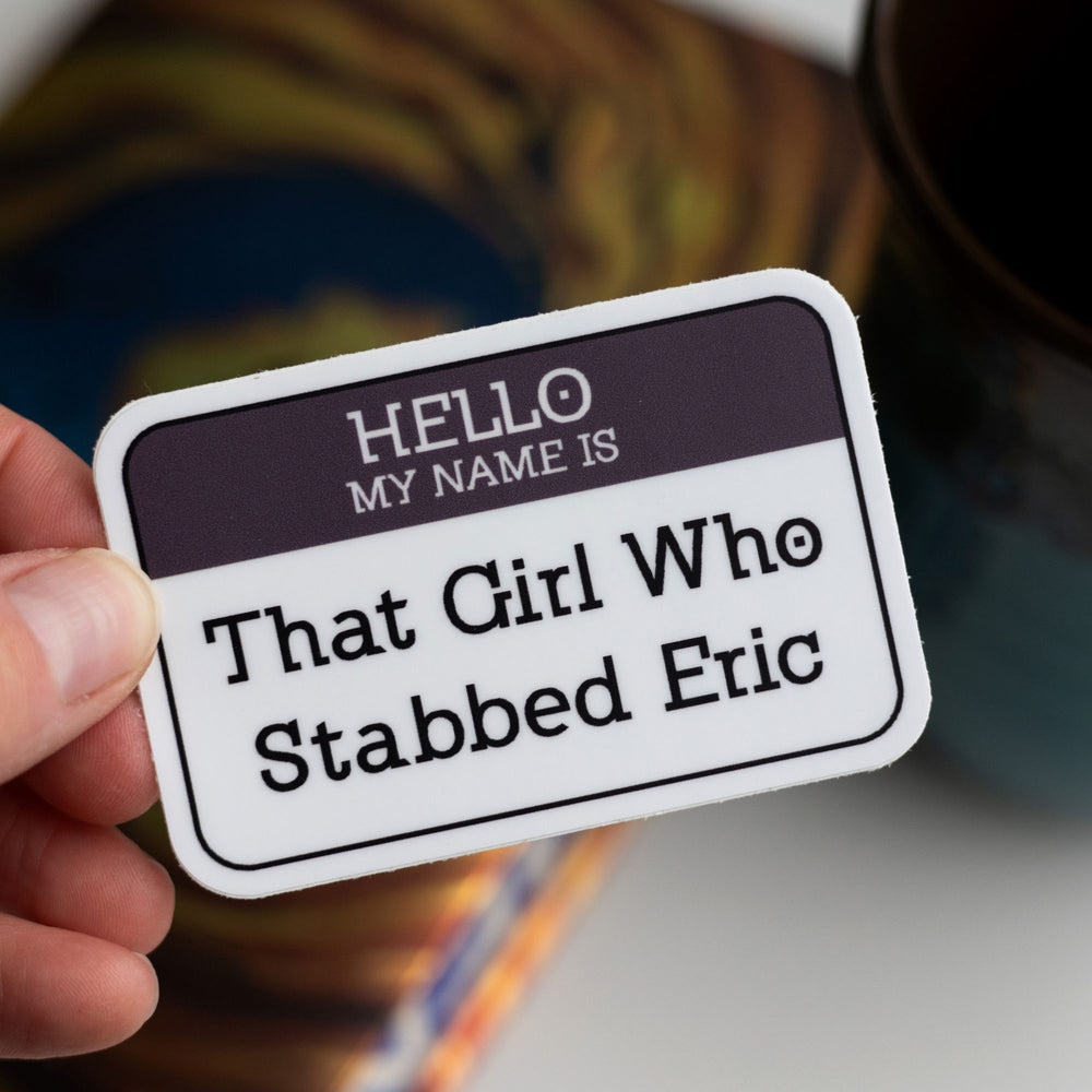Sticker of a ‘Hello my name is…’ nametag with a purple top and ‘That Girl Who Stabbed Eric’ in the white space, held over Divergent by Veronica Roth.
