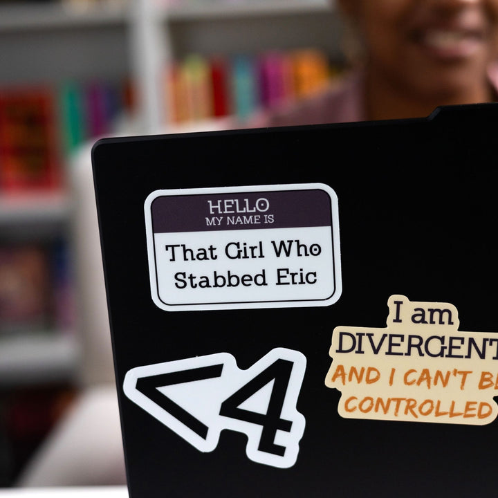 Sticker of a ‘Hello my name is…’ nametag with a purple top and ‘That Girl Who Stabbed Eric’ in the white space, on a black laptop. From Divergent.