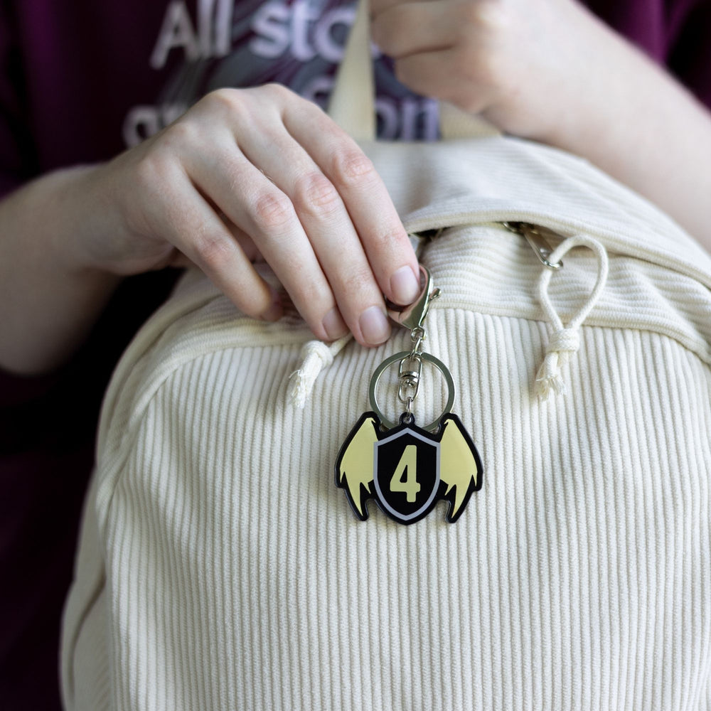  Acrylic keychain of a black shield with golden wings & a gold four on the shield clipped to a white backpack.

